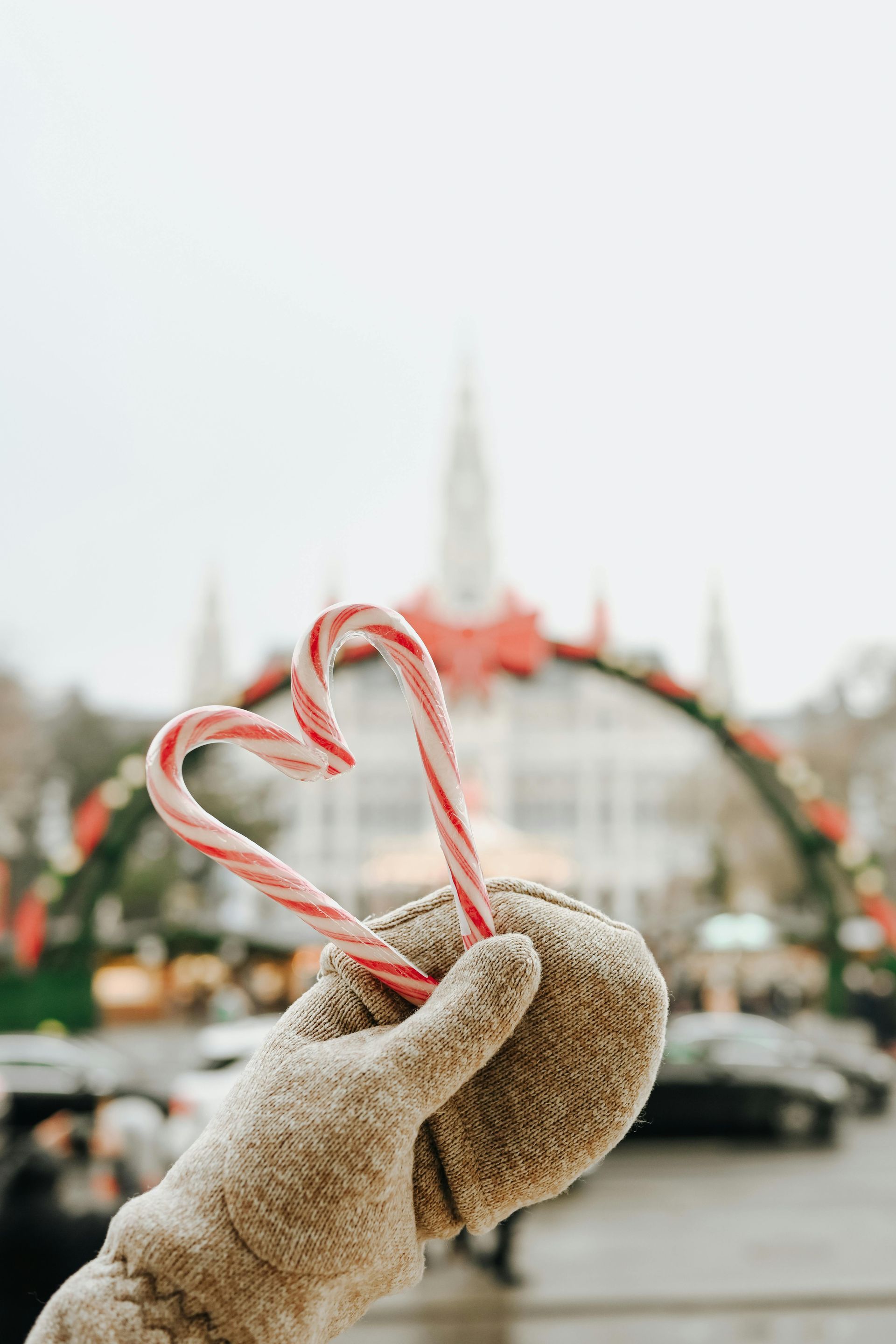 Hand in a mitten holding candy canes shaped into a heart, with a festive arch in the background.