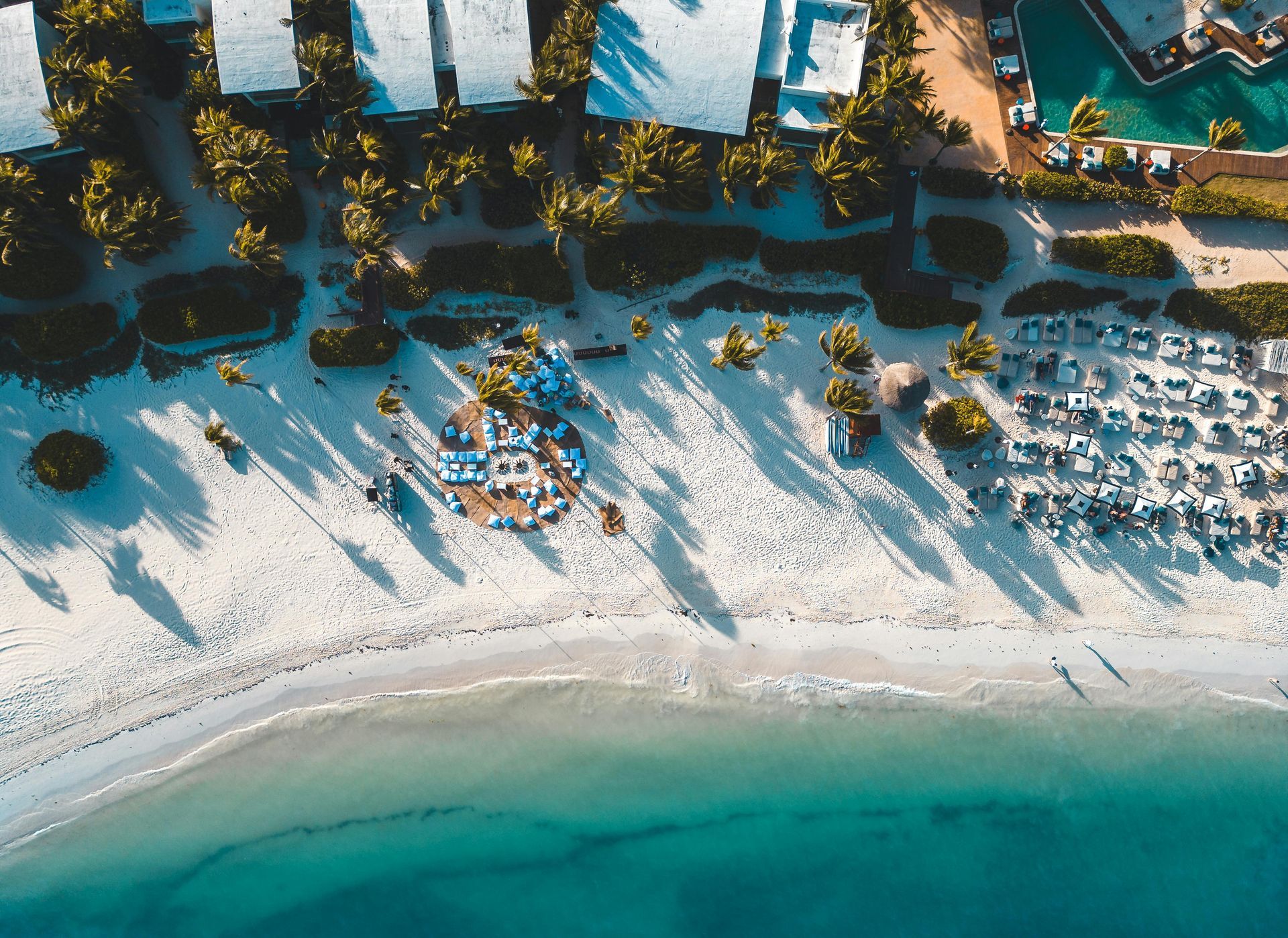 Aerial view of white sand beach, turquoise water, and resort buildings with palm trees casting long shadows.