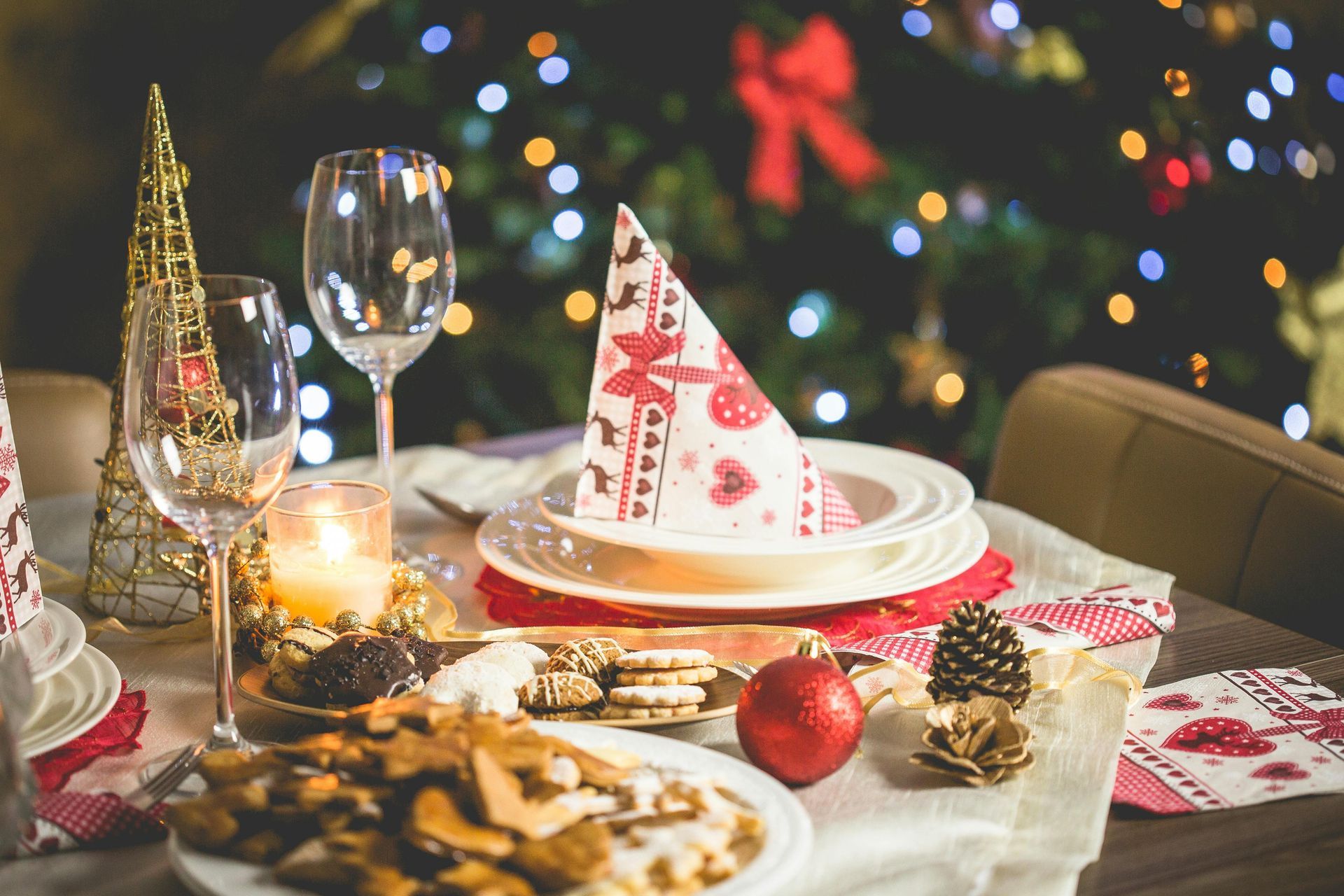Christmas table setting with dishes, wine glasses, and decorations against a blurred tree with lights.