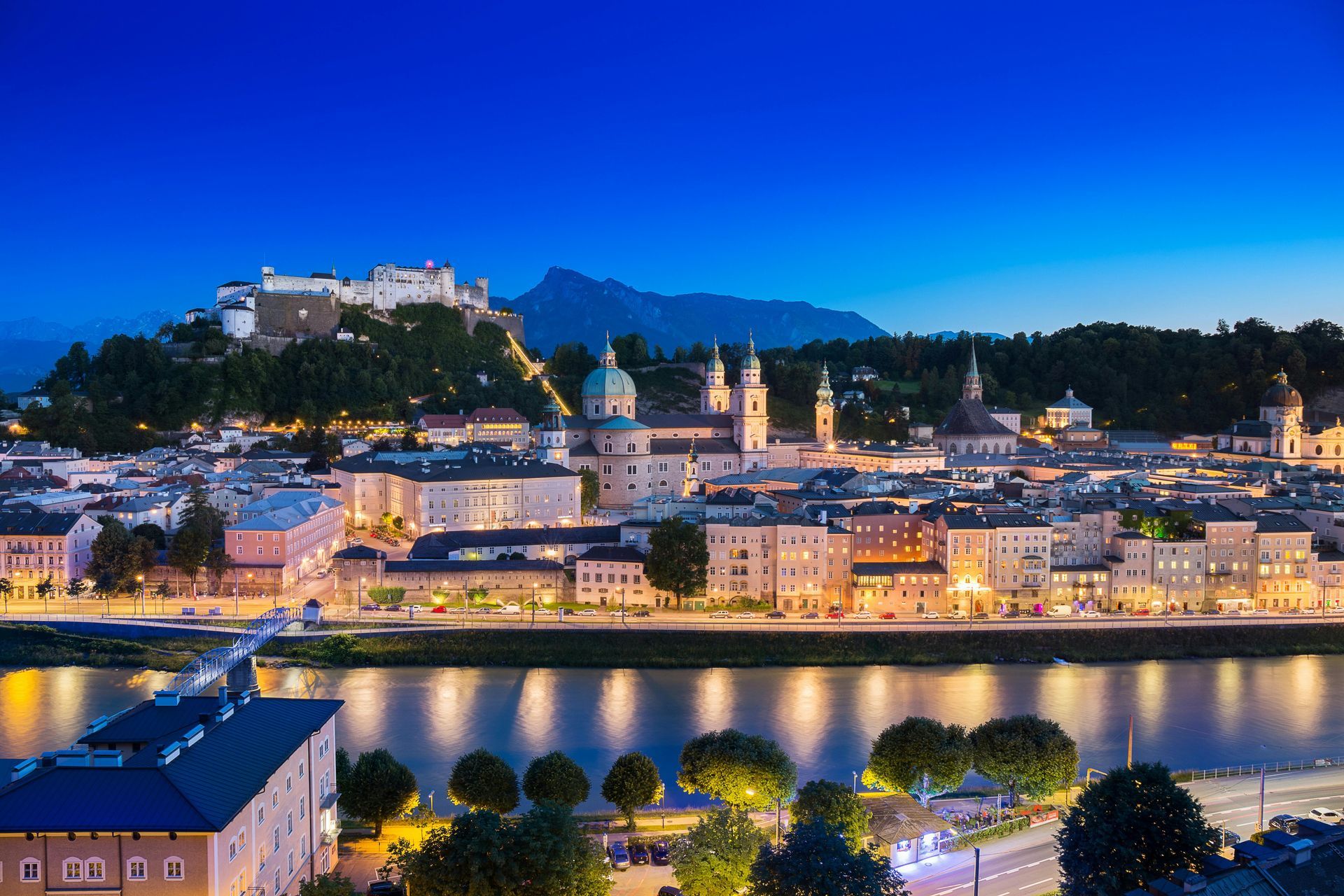 Cityscape at dusk with a river, buildings, and a hilltop castle under a blue sky.
