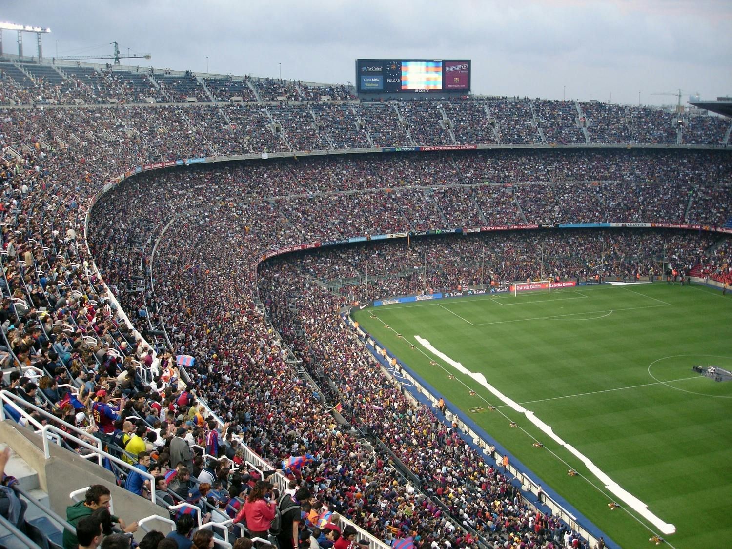 Large, crowded stadium with a green field. Spectators fill the tiered seating under an overcast sky.