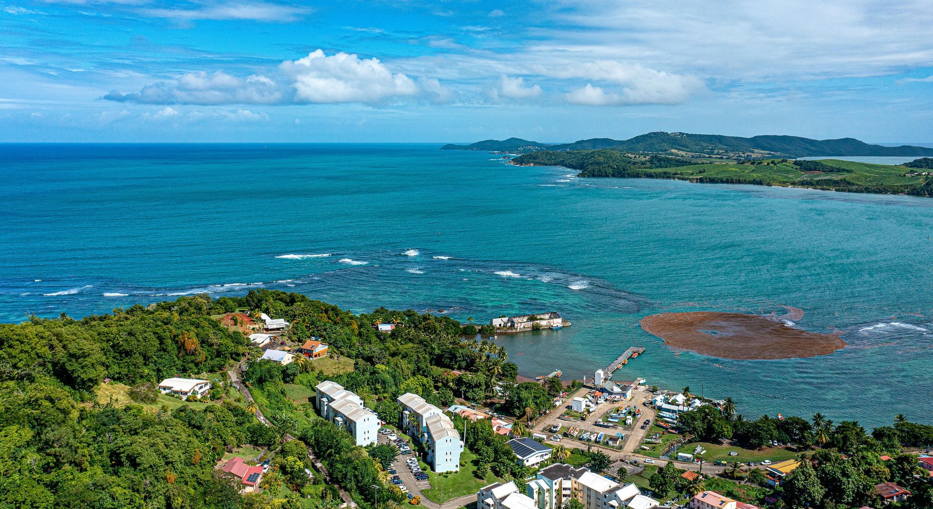 Aerial view of a coastal town with buildings, a dock, and the turquoise ocean meeting the shoreline.