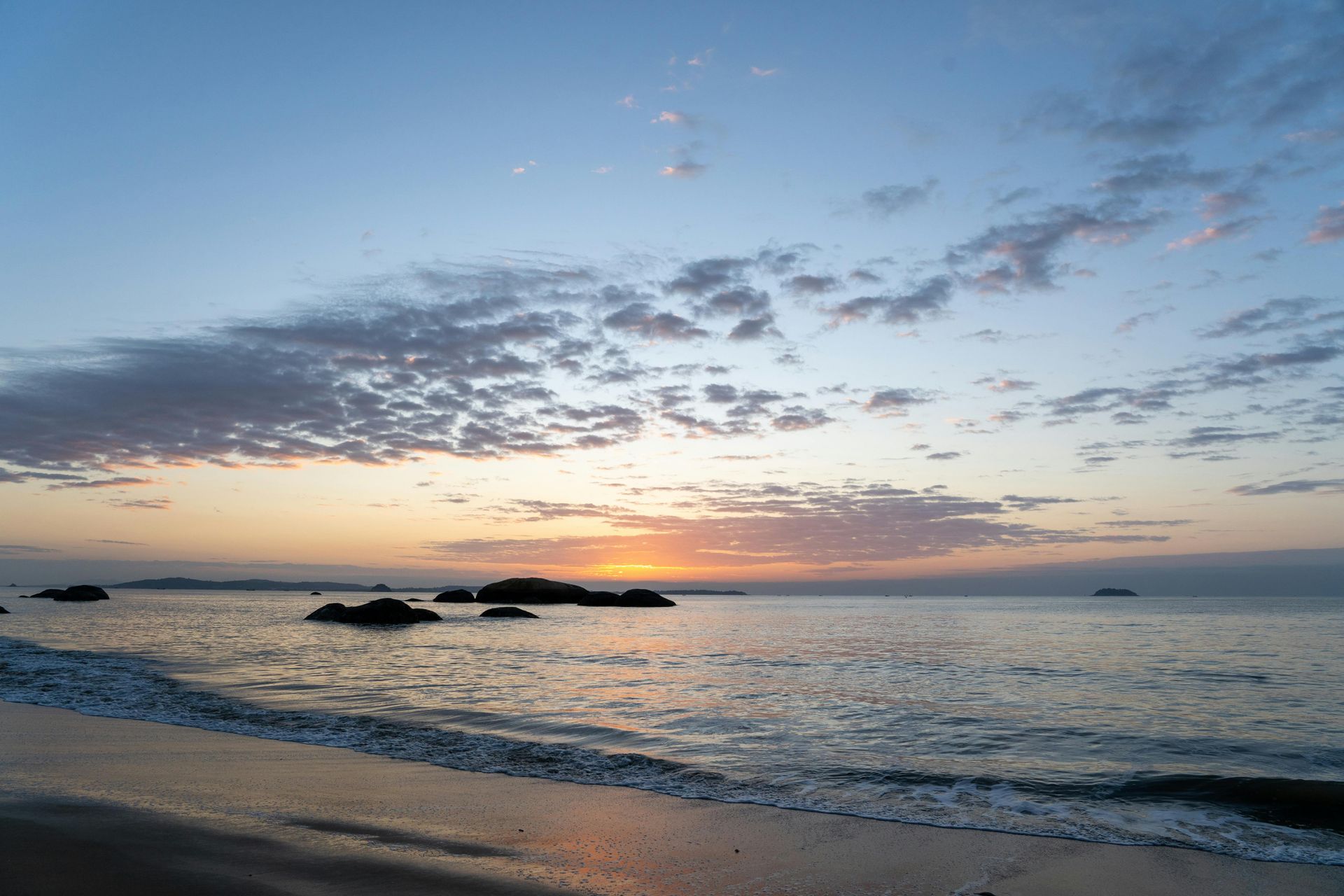 Sunset over the ocean with dark rocks. Sky is blue with orange and pink clouds. Waves lap the shore.