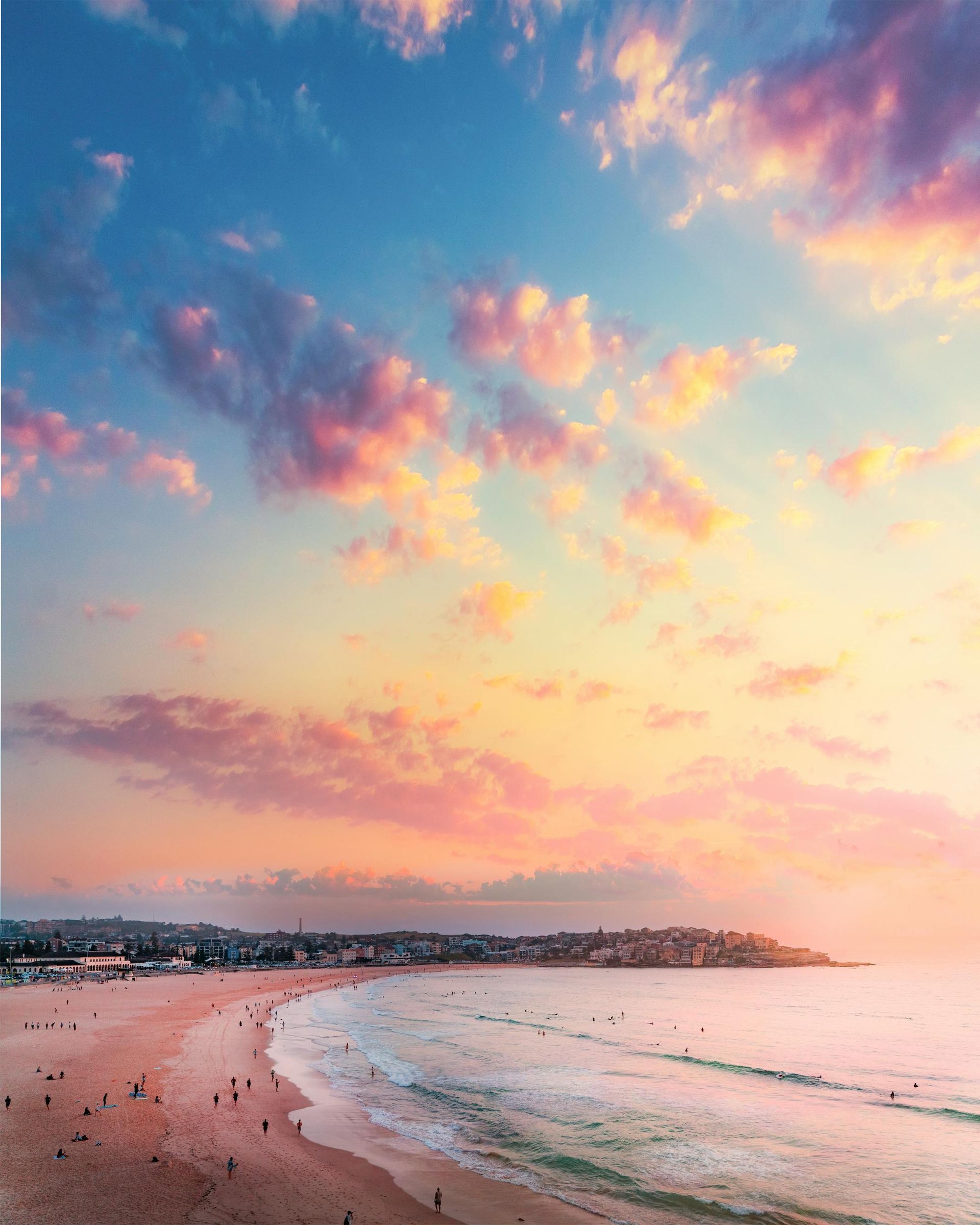 Beach at sunset, with pink and blue sky, waves, and distant buildings.