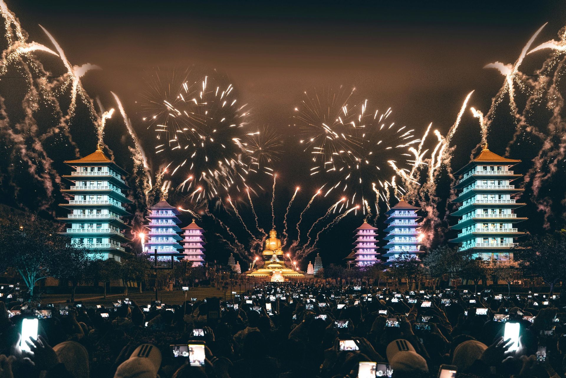 Fireworks over illuminated Asian-style pagodas and a statue, with a crowd of people holding up phones.
