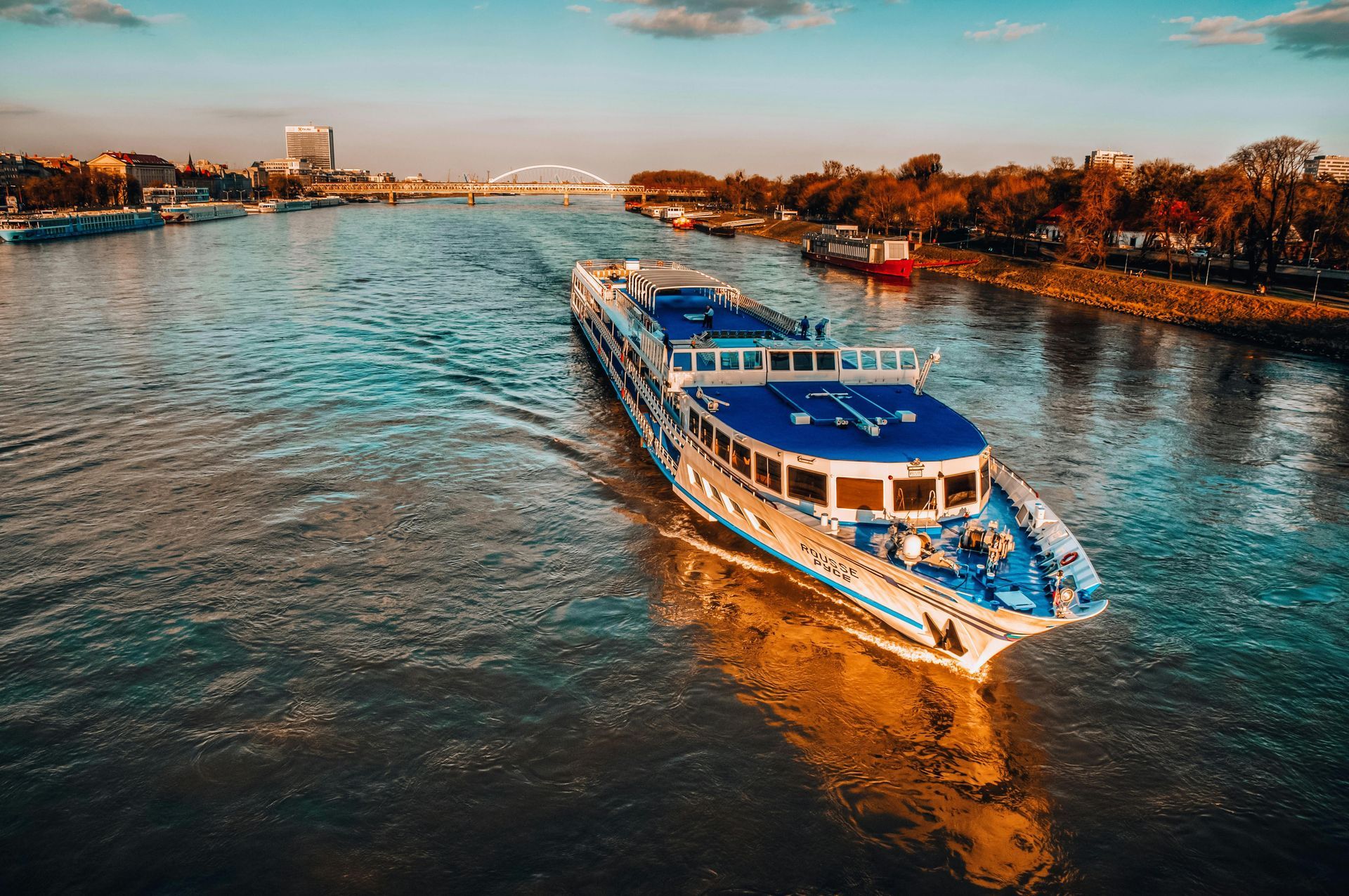 A blue and white passenger river boat travels along a calm river towards a distant bridge under a clear, warm sky.