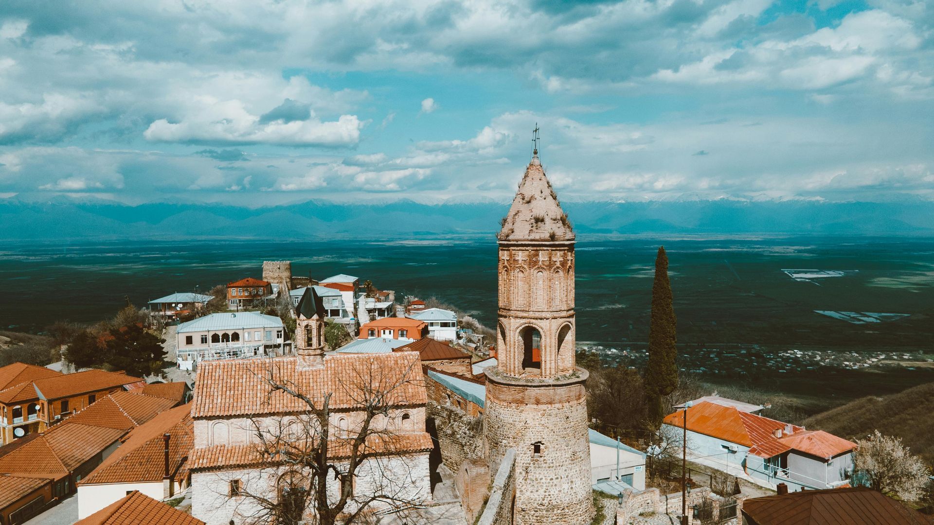 A stone church tower in a town on a hill, with orange roofs and a cloudy sky.
