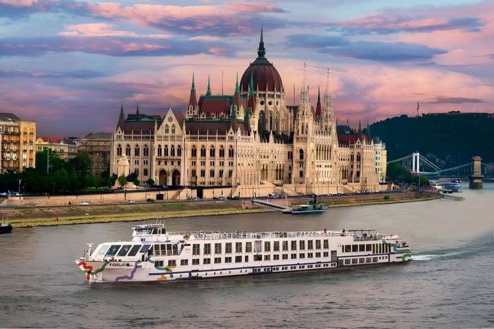 River cruise ship sails past the Hungarian Parliament Building on the Danube River at sunset.