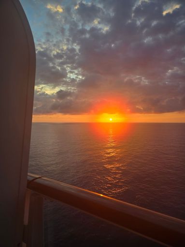 Bright orange sunset over the ocean, viewed from the balcony of a ship with a railing in the foreground.