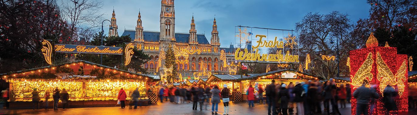 A bustling Christmas market in front of a grand illuminated building. Many people are walking through the market, lit up by twinkling lights.