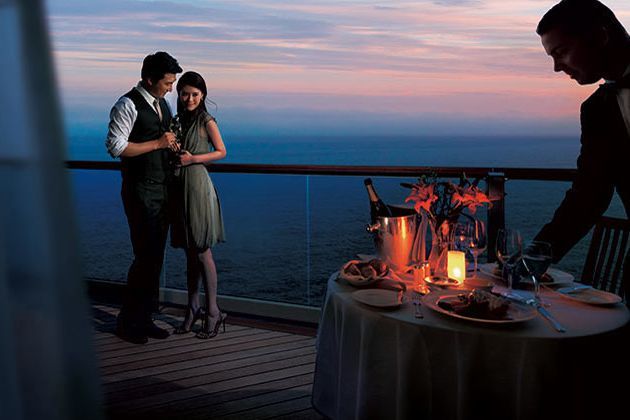 Couple enjoying champagne dinner on a balcony overlooking the ocean at sunset. A waiter stands nearby.