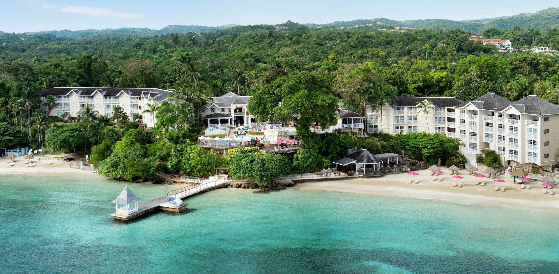 Beachfront resort with white buildings, clear turquoise water, and lush green trees.