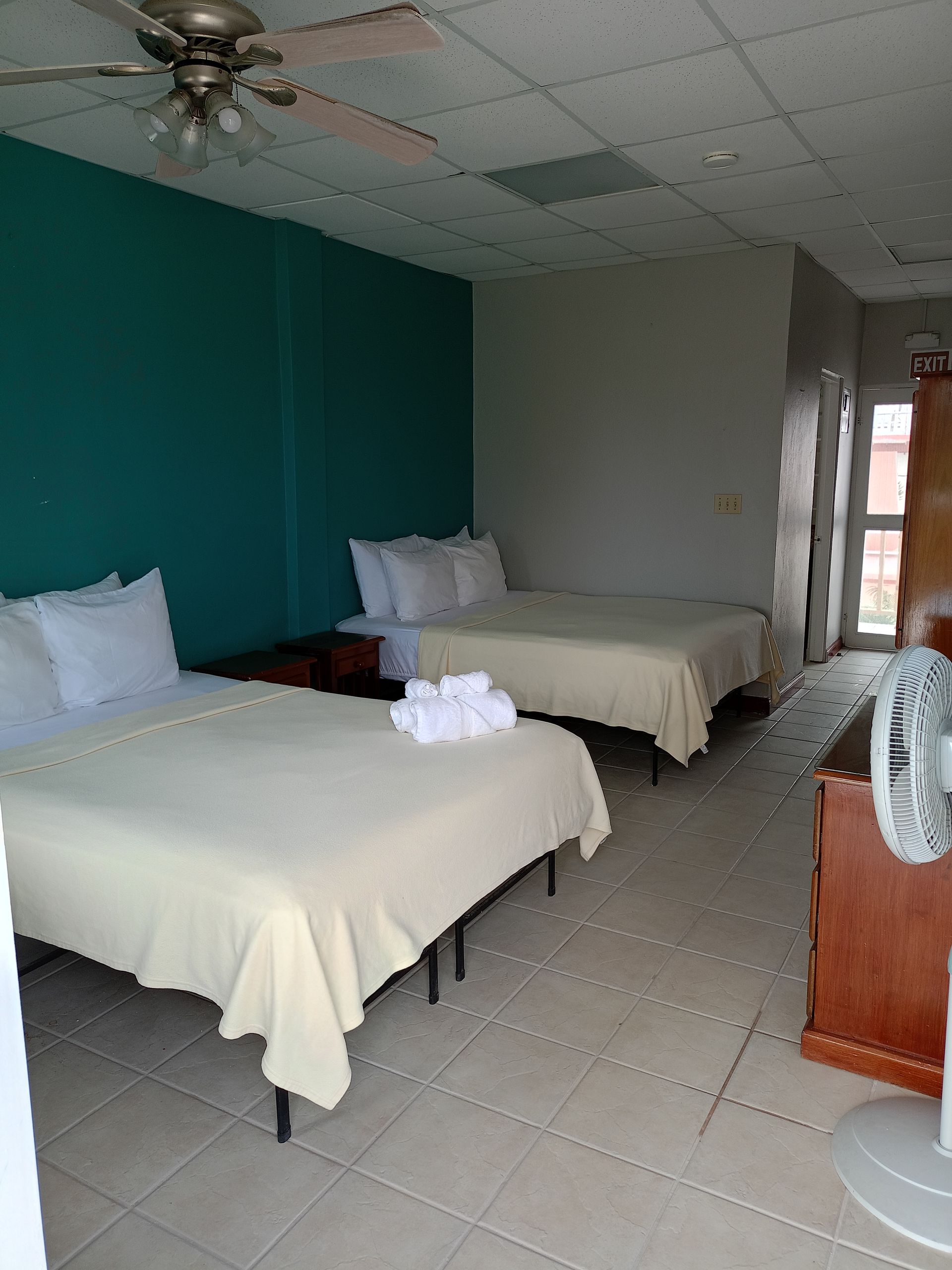 Two beds in a room, teal wall, off-white linens, towels on a bed, fan in the corner.