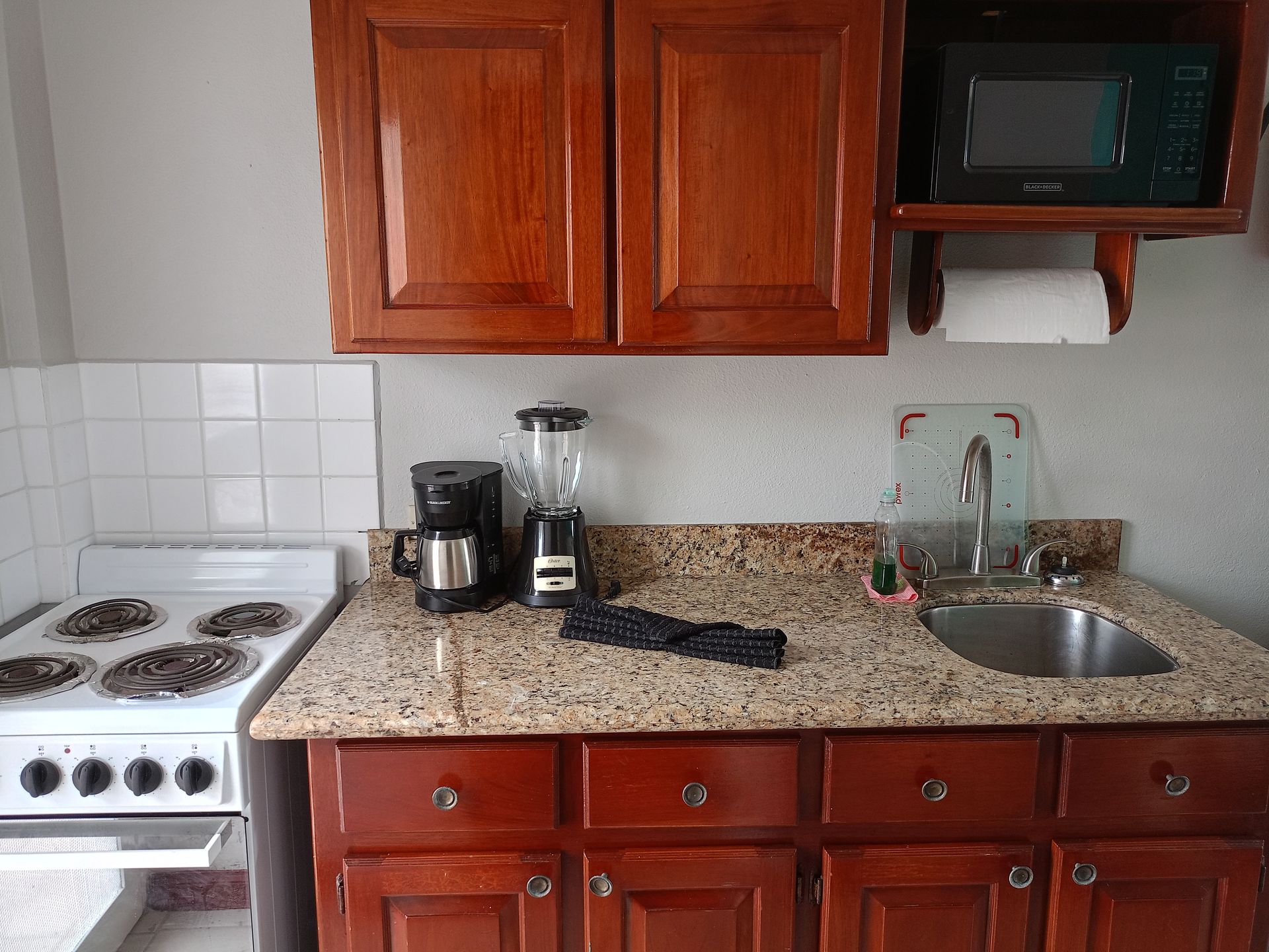 Small kitchen with brown cabinets, granite countertop, stove, and sink. Includes microwave, blender, and coffee maker.