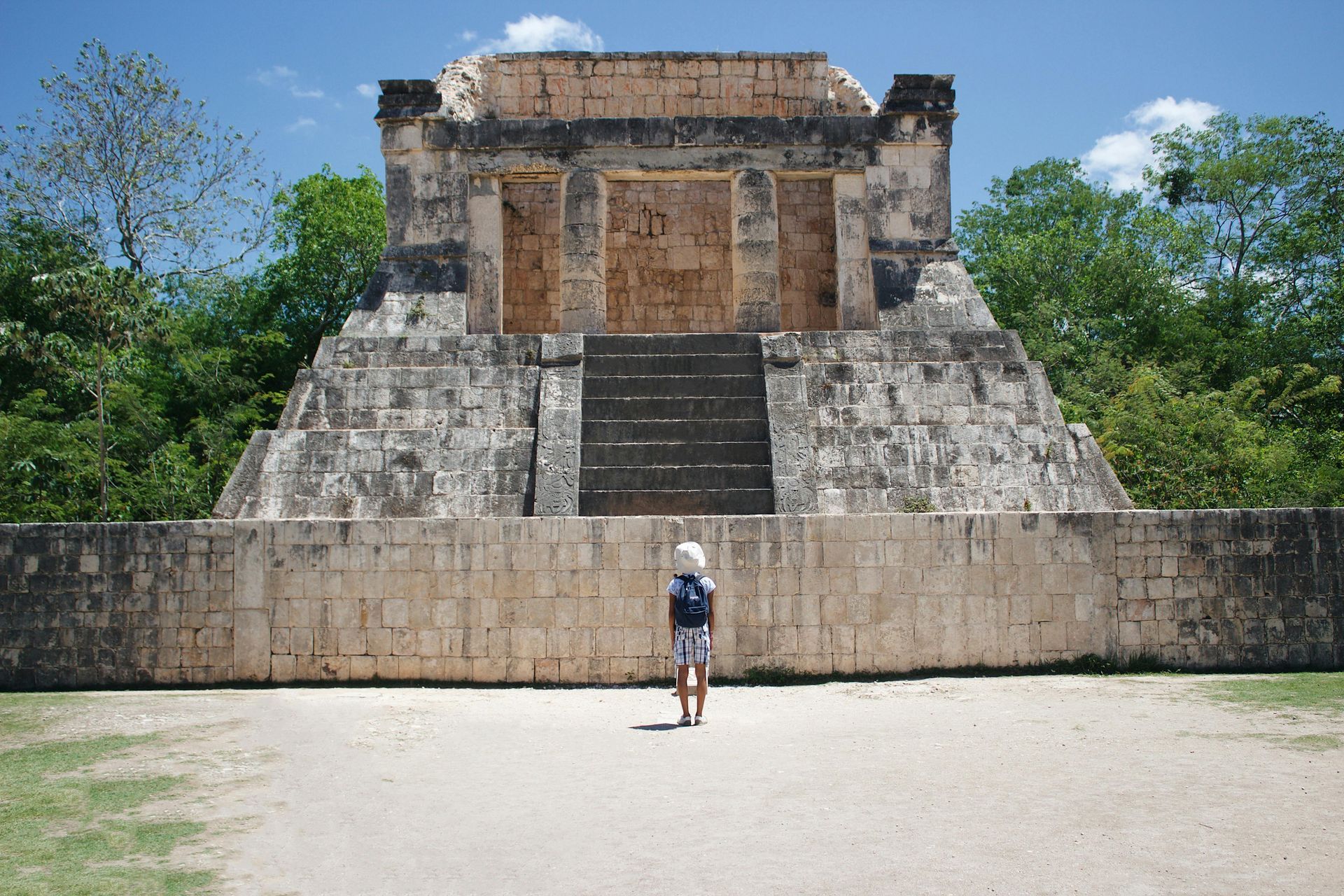 Person facing a stone Mayan temple with a large open plaza and lush trees in the background.