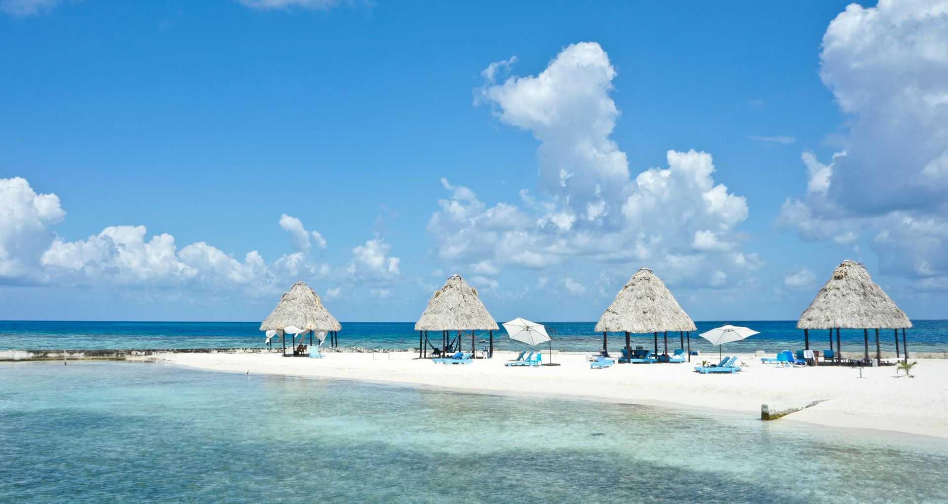 White sand beach with thatched-roof cabanas, clear blue water, and partly cloudy sky.