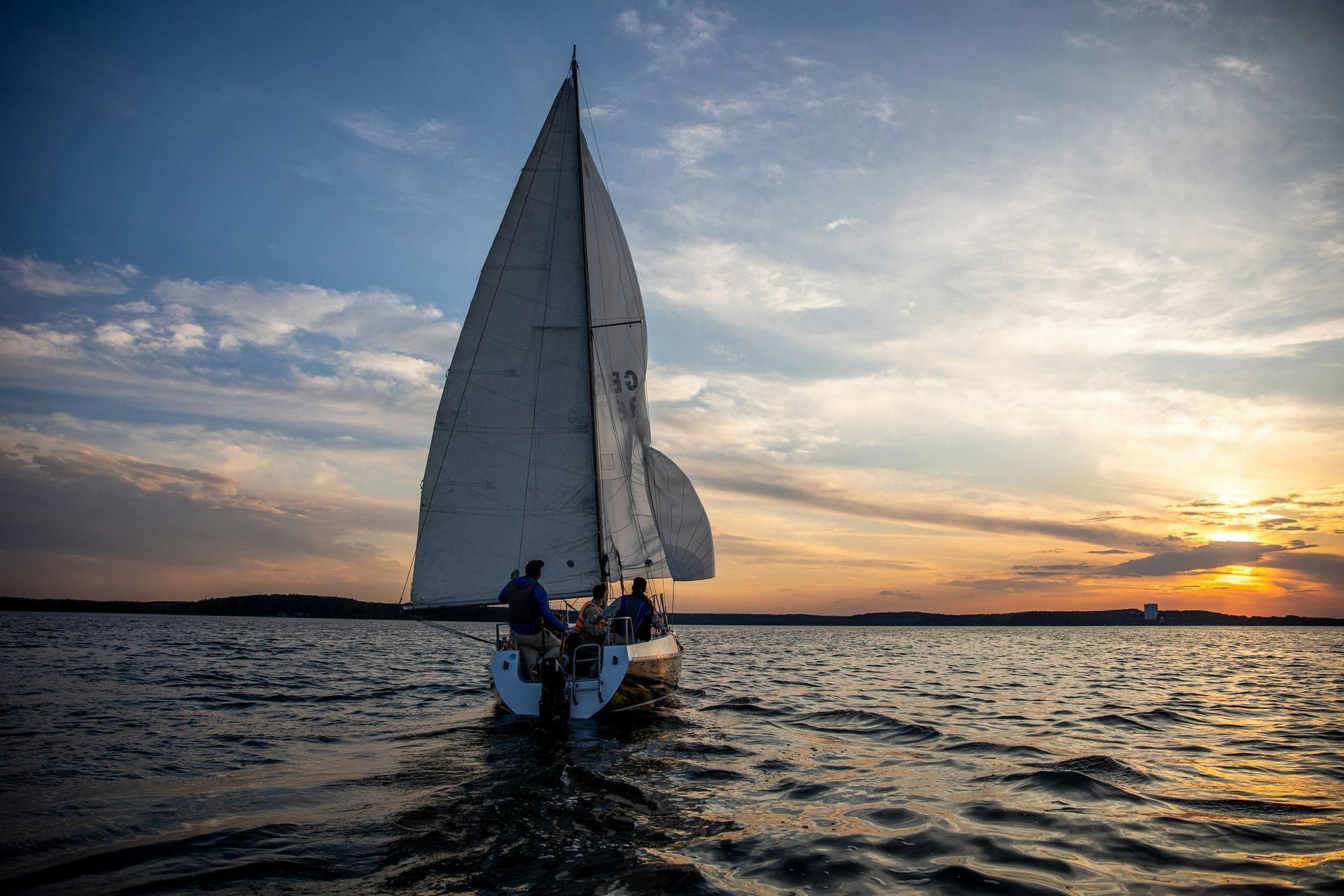 Sailboat on water at sunset, white sail billowing, several people visible on deck.