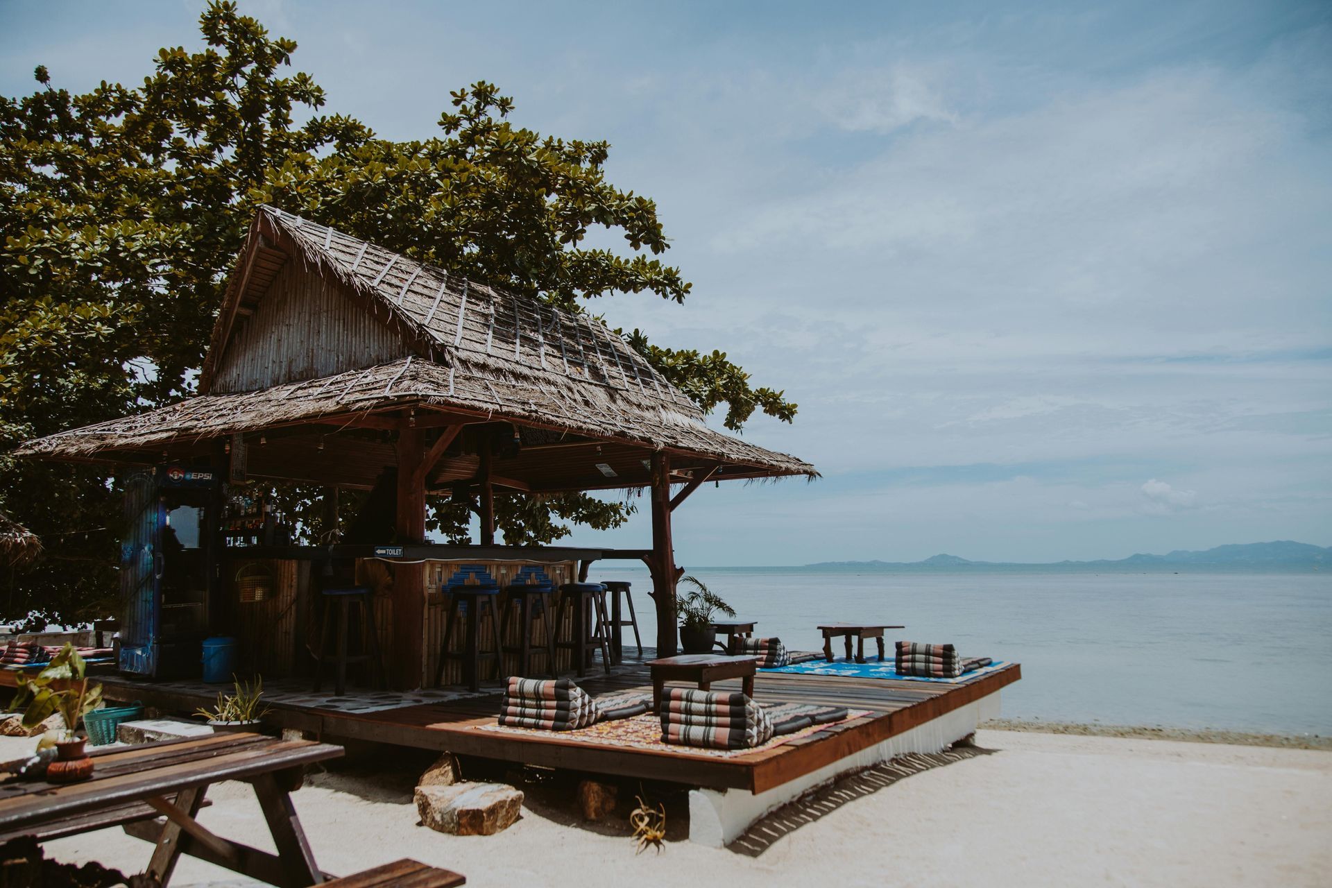 Beach bar with thatched roof overlooking ocean, blue sky, white sand.