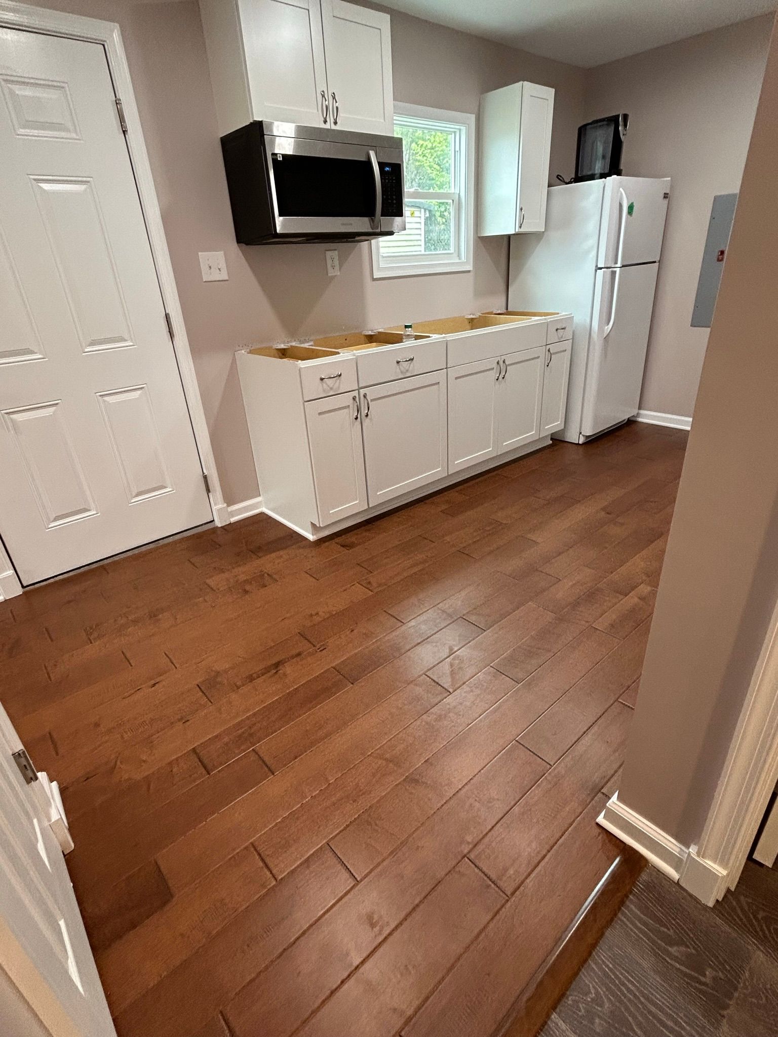 A kitchen with hardwood floors , white cabinets , a refrigerator and a microwave.