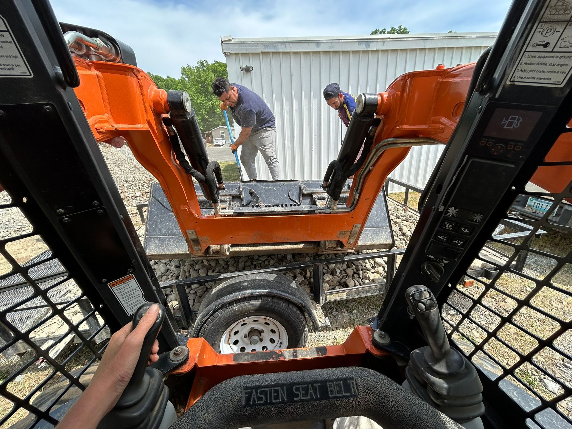 A person is driving a skid steer with a man standing behind it.