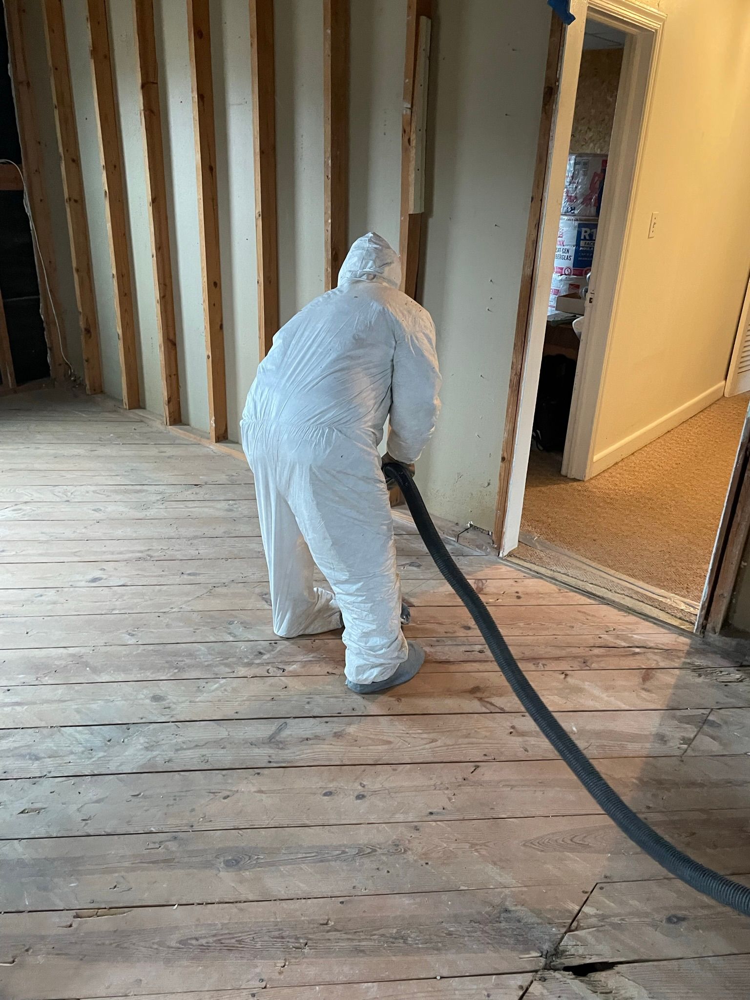 A man in a protective suit is using a vacuum hose to clean a wooden floor.