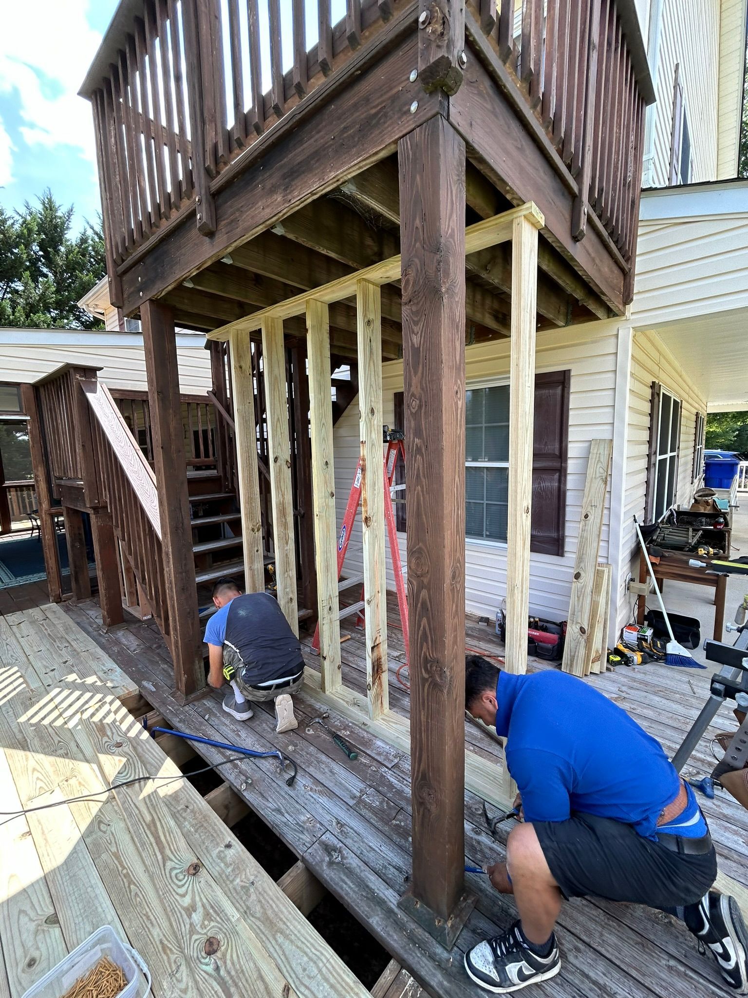 A man is kneeling on a wooden deck under a deck being built.