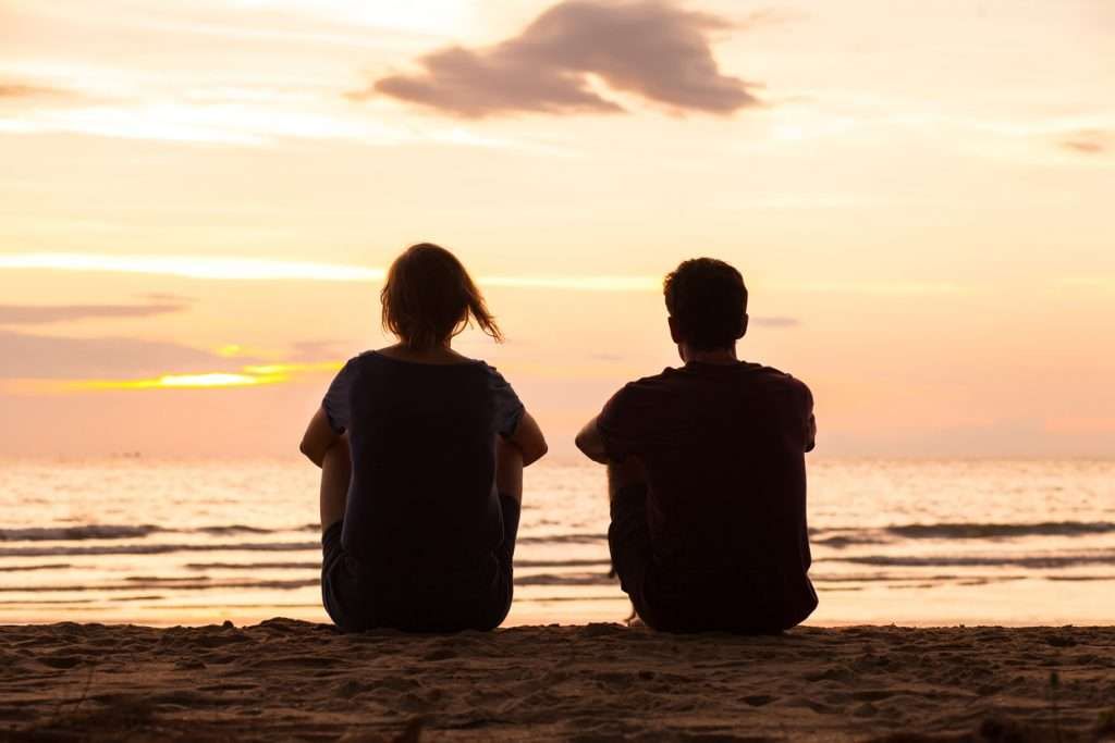Two people sit on a beach, silhouetted against a sunset over the ocean.