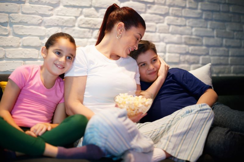 Woman with children watching something on a couch with popcorn.