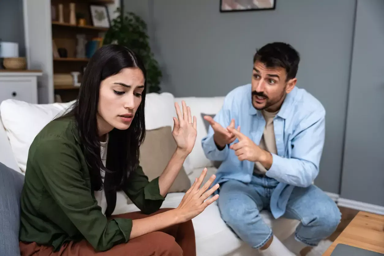 Woman with hand raised, trying to stop a man pointing and speaking while sitting on a couch.