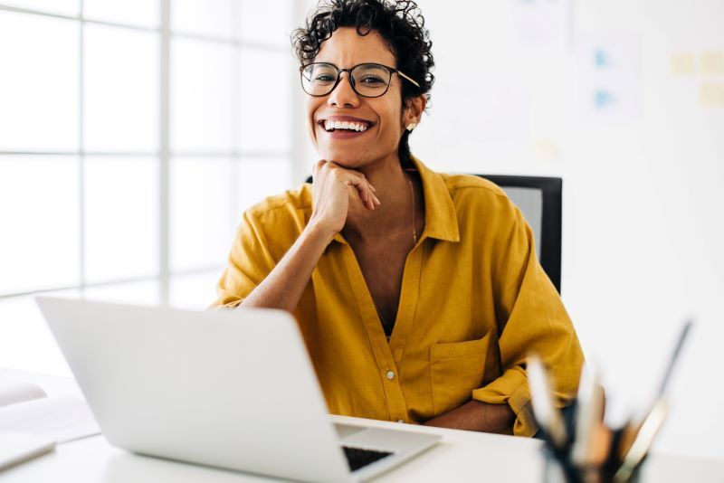 Woman wearing glasses, smiling, sitting at a desk with a laptop, hand on chin, indoors.
