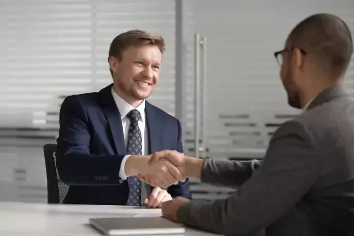 Two men in suits shaking hands across a table, smiling.