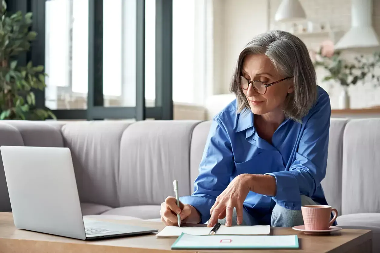 Woman with gray hair and glasses, writing in notebook while looking at laptop. Sitting on couch, indoors.