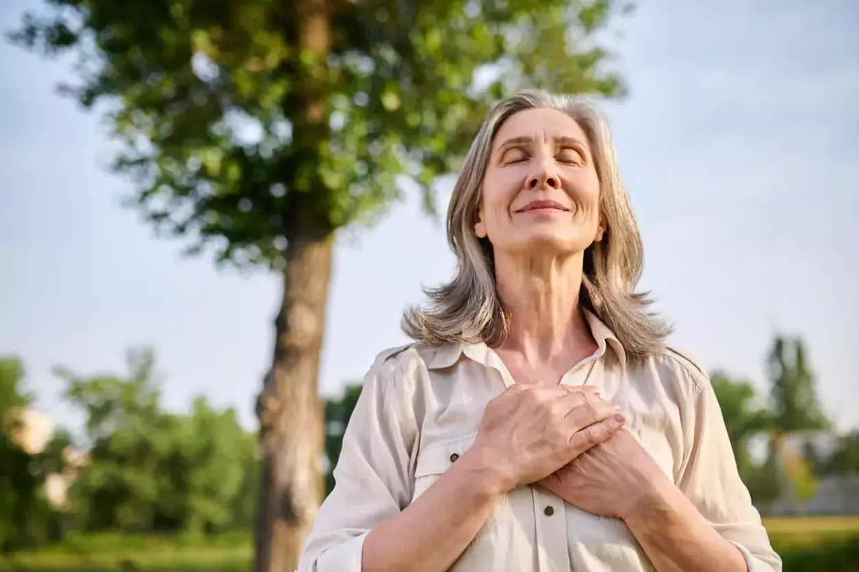Woman with eyes closed, hands on chest, smiling gently outdoors.