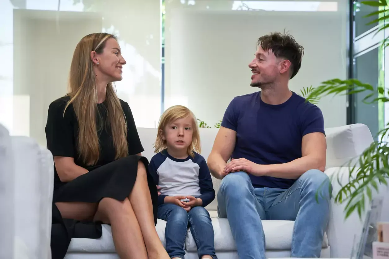 Family of three smiling while sitting on a white sofa in a waiting room.