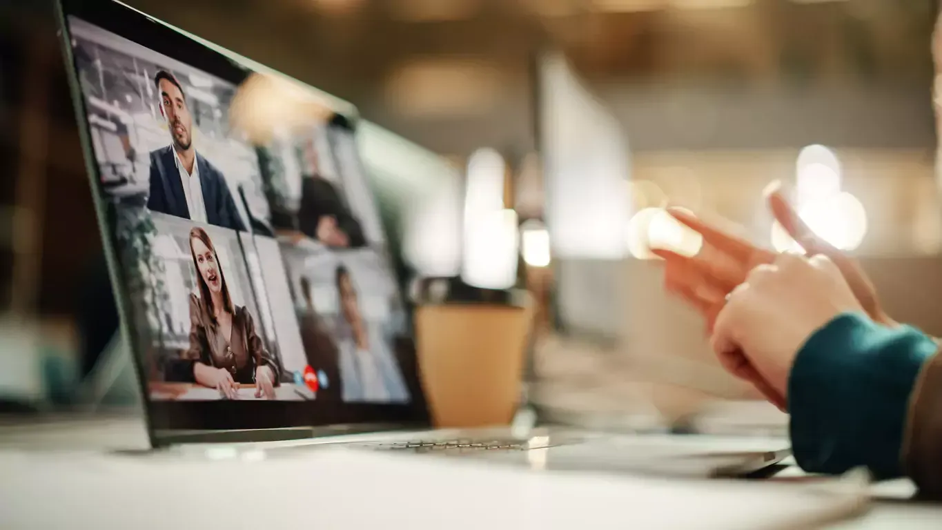 Laptop screen displaying a video call with four participants. Someone's hands gesture near the screen.