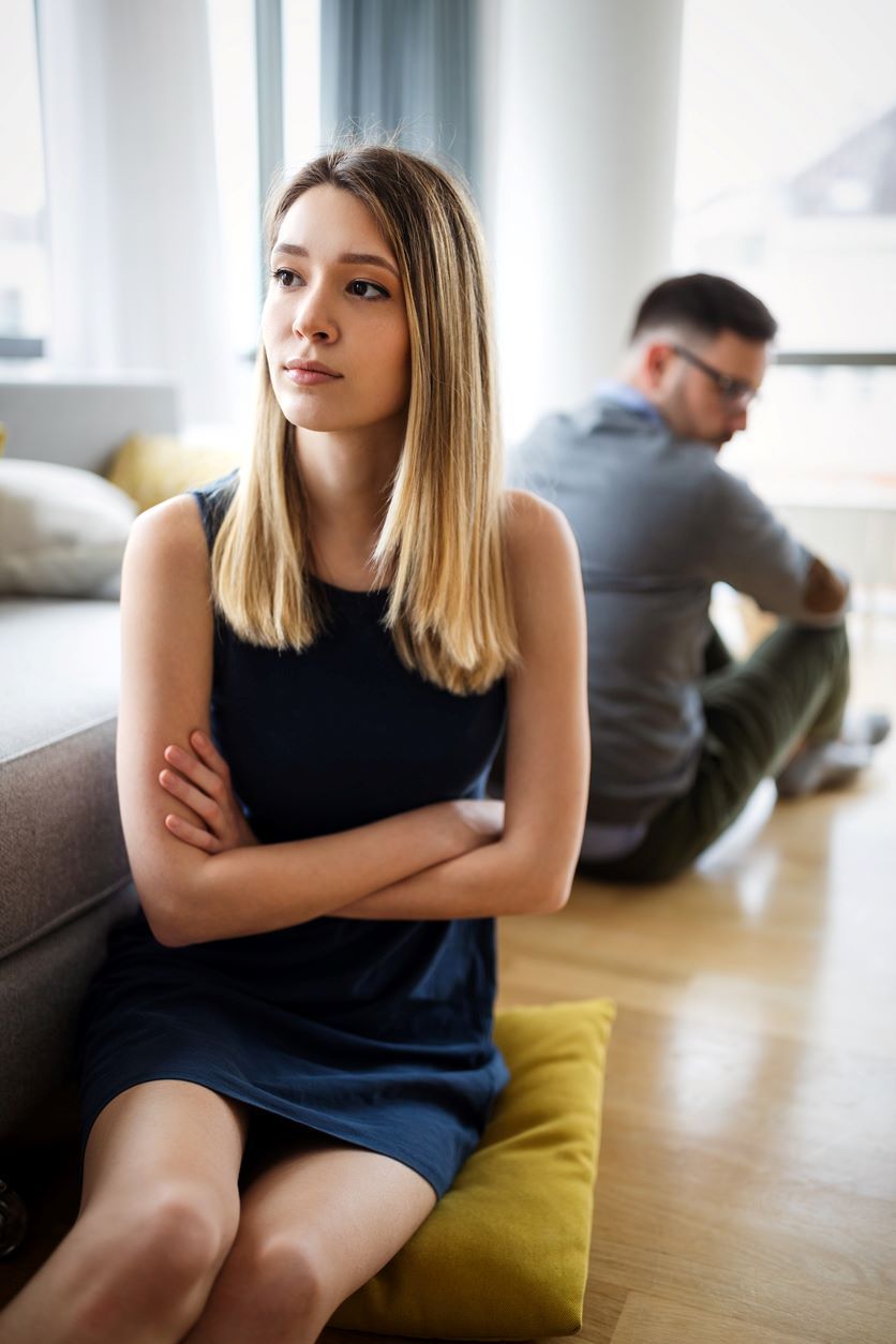 Woman sits on end of sofa looking away from man who appears upset - decorative image