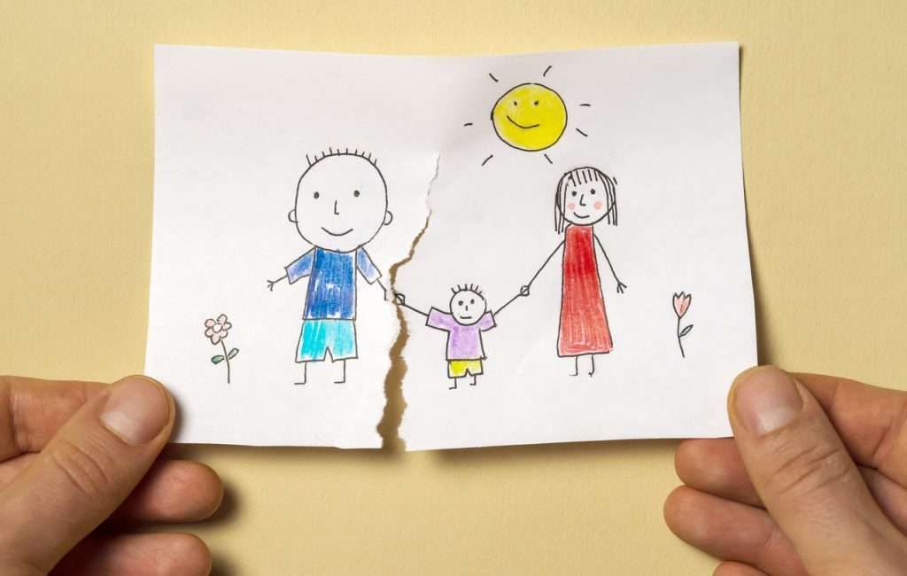 Hands tearing a drawing of a family in half. The drawing shows a father, child, and mother under a sun, with flowers.