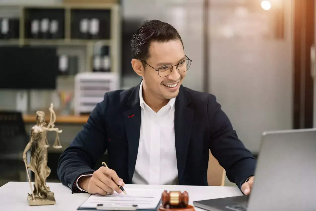 Man in suit smiles while using laptop, pen in hand, in office setting, statue of justice and gavel present.