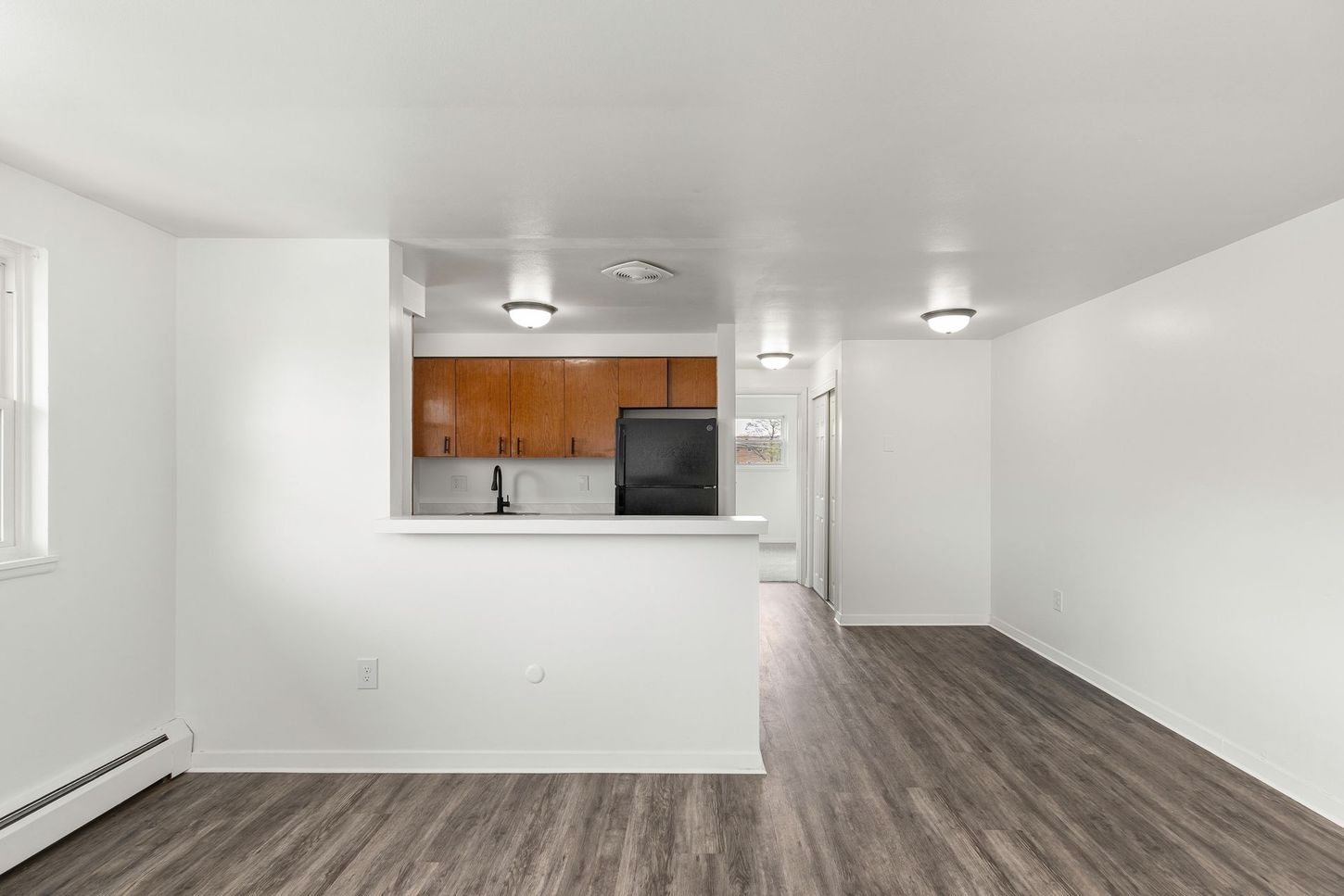 Empty living space with kitchen bar, wood cabinets, black appliances, and grey wood-look flooring.