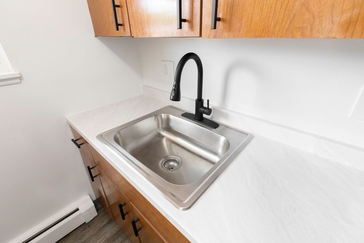 Kitchen sink with black faucet, stainless steel sink, white countertop, and wood cabinets.