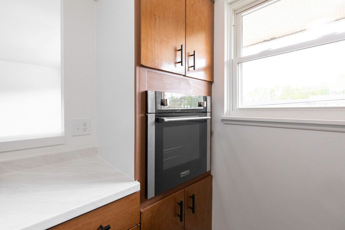Built-in oven surrounded by wooden cabinets in a kitchen. Next to a window.