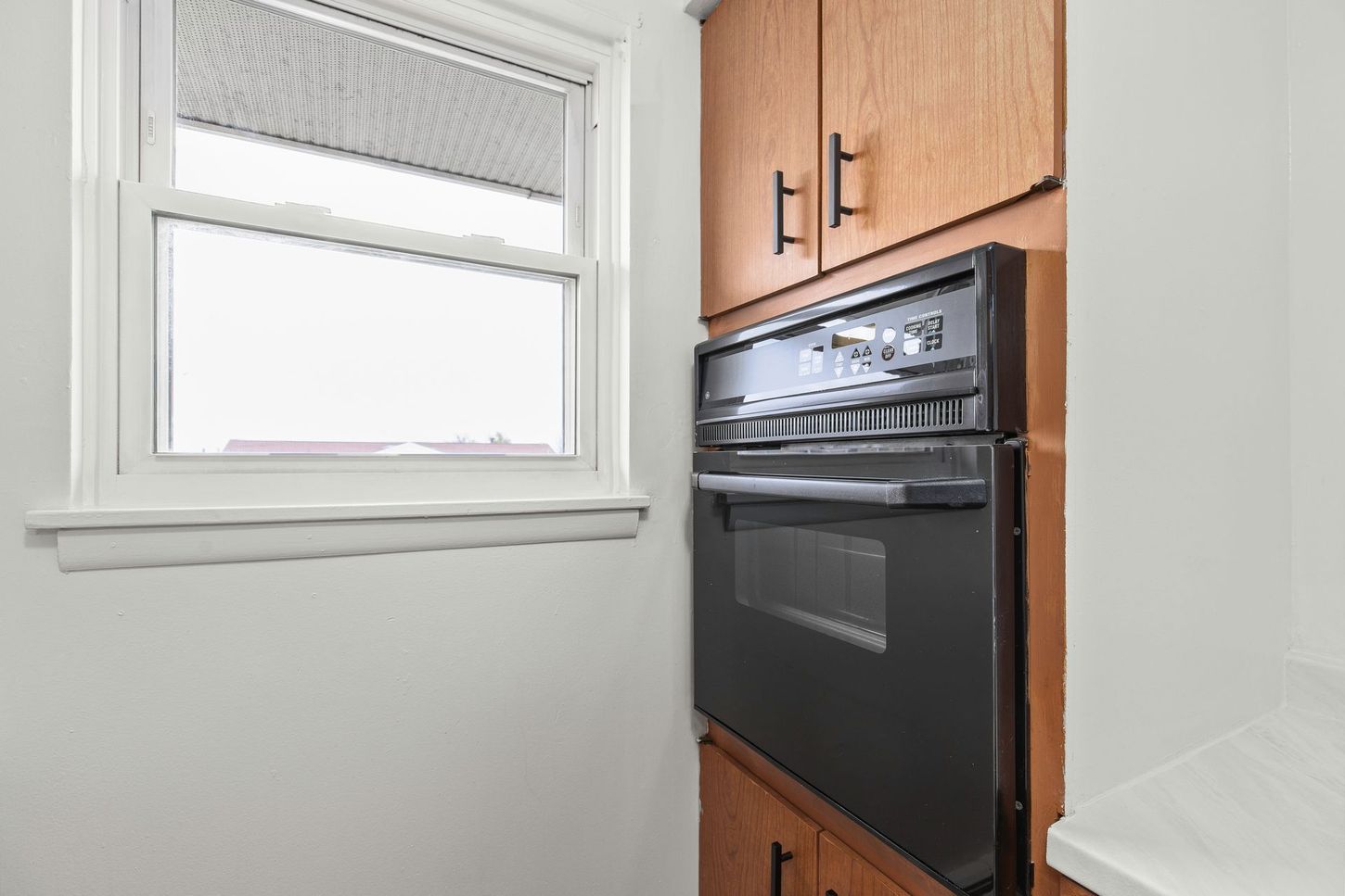 Kitchen with a built-in black oven next to a window with a closed blind, surrounded by wood cabinetry.