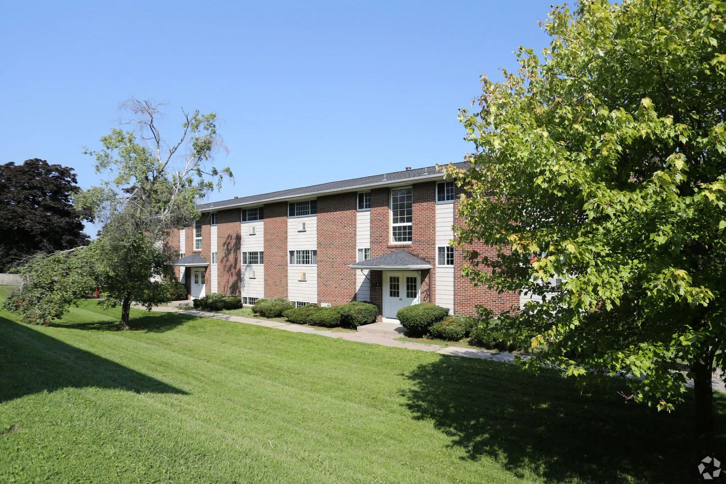 Two-story brick apartment building with green lawn, trees, and a blue sky.