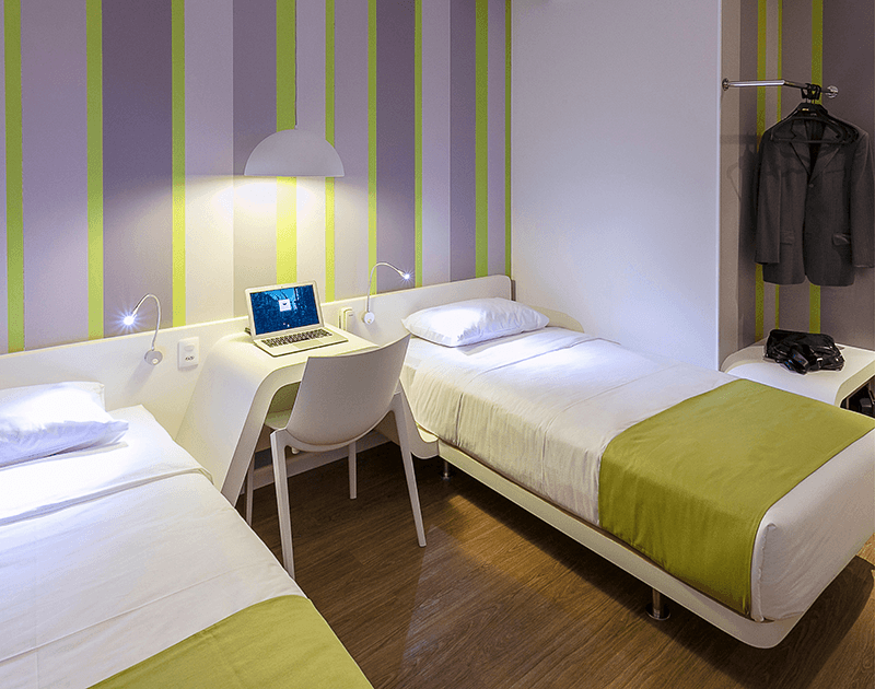 Um quarto de hotel bem iluminado com duas camas de solteiro, uma escrivaninha, um laptop e papel de parede listrado em roxo e verde limão.