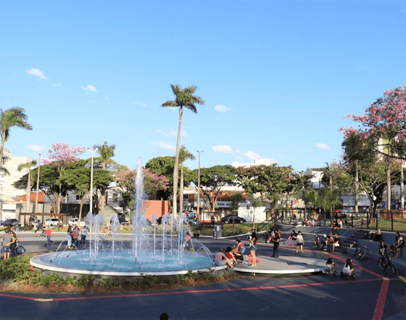Uma fonte circular jorra água para cima em uma praça pública ensolarada, cercada por palmeiras e pessoas relaxando.