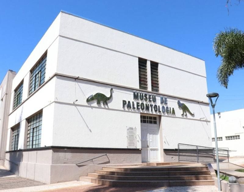 Um edifício branco de dois andares do Museu de Paleontologia sob um céu azul claro, apresentando silhuetas de dois dinossauros.