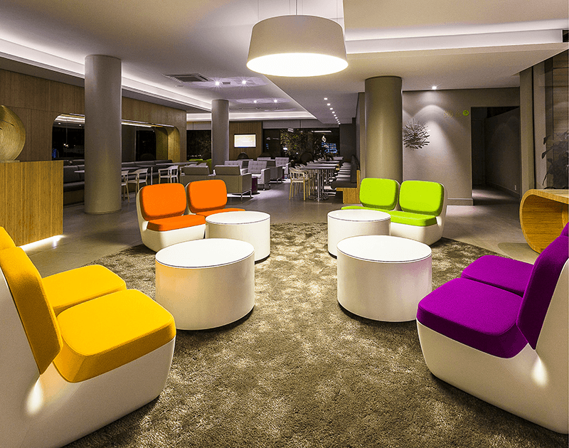 Um lobby de hotel moderno com assentos modulares coloridos em tons de laranja, amarelo, verde limão e roxo, dispostos sobre um tapete.