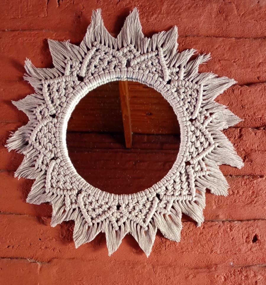 Un espejo circular con un marco de macramé blanco con efecto sol contra una pared de ladrillos rojos.