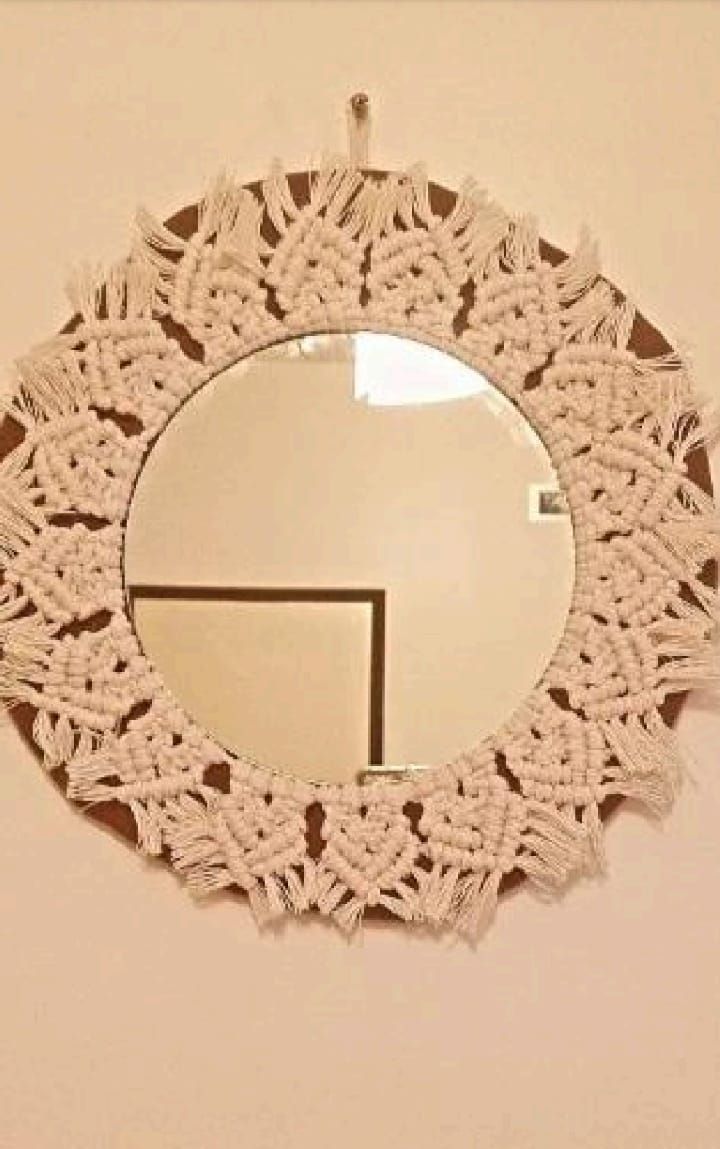 Un espejo circular con un marco de macramé blanco con efecto sol contra una pared de ladrillos rojos.
