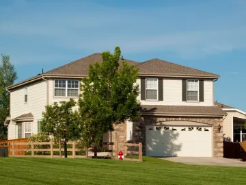 Two-story suburban house with beige siding, brown roof, and attached two-car garage behind a wooden fence.